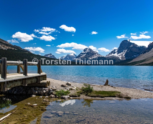 Lakeview-1 Bergsee in der Nähe von Banff in Kanada. Berge im Hintergrund. Blauer Himmel mit weißen Wolken. Holzsteg. Seine im Wasser zu erkennen.