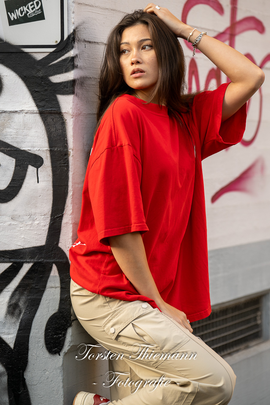Modeaufnahmen einer jungen, asiatisch wirkenden Frau mit rotem Shirt und langen dunklen Haaren, vor einer Wand mit Graffiti.