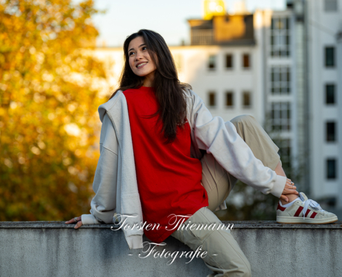 Modeaufnahmen einer jungen, asiatisch wirkenden Frau mit rotem Shirt und langen dunklen Haaren, sitzend auf einer Mauer.