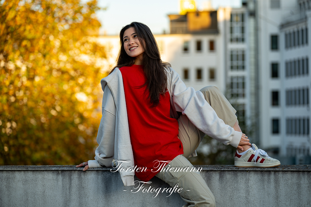 Modeaufnahmen einer jungen, asiatisch wirkenden Frau mit rotem Shirt und langen dunklen Haaren, sitzend auf einer Mauer.