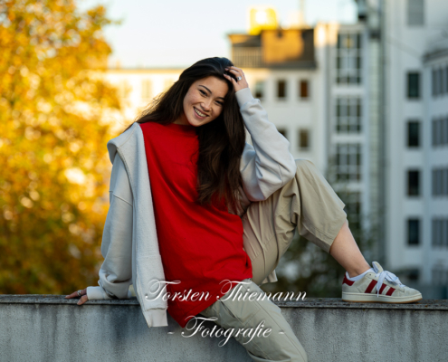 Modeaufnahmen einer jungen, asiatisch wirkenden Frau mit rotem Shirt und langen dunklen Haaren, sitzend auf einer Mauer.