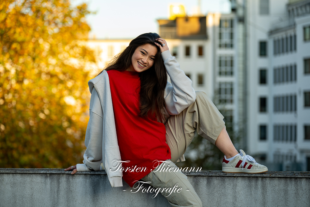 Modeaufnahmen einer jungen, asiatisch wirkenden Frau mit rotem Shirt und langen dunklen Haaren, sitzend auf einer Mauer.