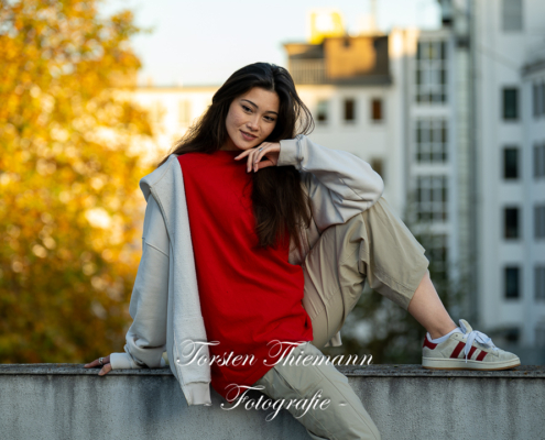 Modeaufnahmen einer jungen, asiatisch wirkenden Frau mit rotem Shirt und langen dunklen Haaren, sitzend auf einer Mauer.