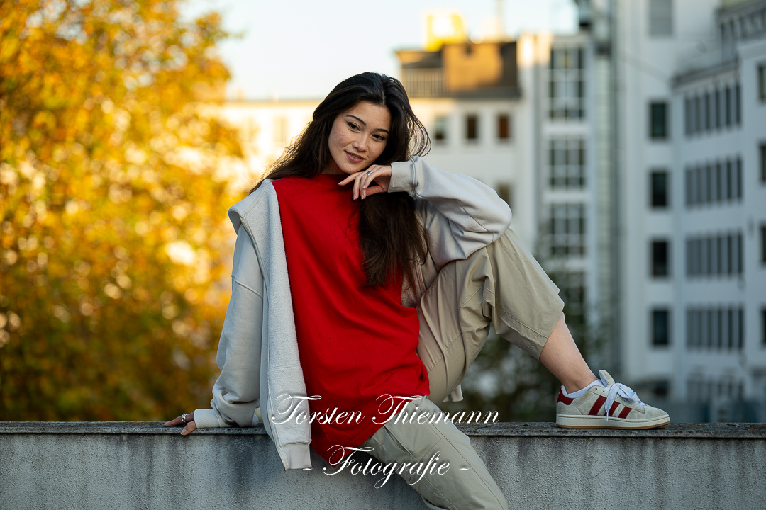 Modeaufnahmen einer jungen, asiatisch wirkenden Frau mit rotem Shirt und langen dunklen Haaren, sitzend auf einer Mauer.